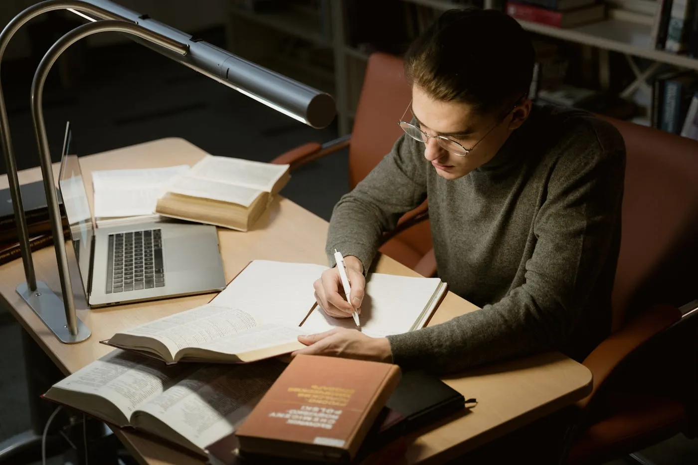 A nursing student studying at a library desk with textbooks, a notebook, and a laptop
